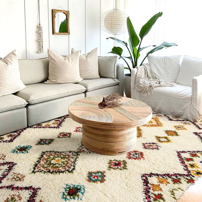 Op de afbeelding: Een wit, pluche vloerkleed met een geometrisch patroon in tinten rood, geel en blauw. Een lichte houten salontafel met een geweven ontwerp staat op het vloerkleed. Een witte bank en een fauteuil staan op de achtergrond.