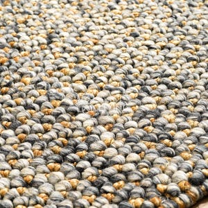 Puede incluir: Una alfombra gruesa tejida a mano con una superficie densa y texturizada hecha de nudos de hilo gruesos y redondeados. La alfombra muestra una paleta de colores variados de gris carb&oacute;n, gris claro, crema y tonos amarillo mostaza c&aacute;lidos.