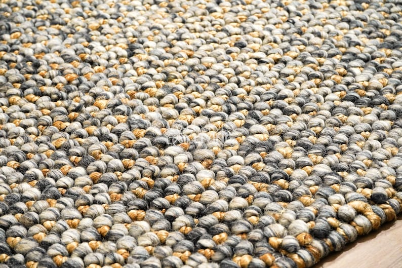 Puede incluir: Una alfombra gruesa tejida a mano con una superficie densa y texturizada hecha de nudos de hilo gruesos y redondeados. La alfombra muestra una paleta de colores variados de gris carb&oacute;n, gris claro, crema y tonos amarillo mostaza c&aacute;lidos.