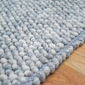 May include: Close-up of a gray and white wool rug with a blue border. The rug is made of small, round balls of wool.