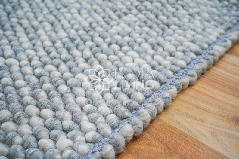 May include: Close-up of a gray and white wool rug with a blue border. The rug is made of small, round balls of wool.