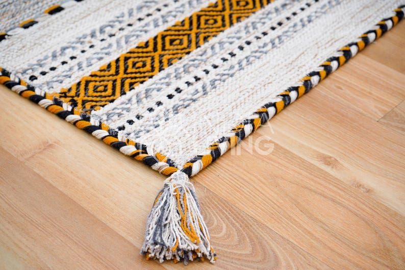 May include: A white, yellow, and black patterned rug with a fringe border and a tassel. The rug is on a wooden floor.