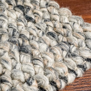 Puede incluir: Primer plano de una alfombra tejida gris y beige con un diseño texturizado en bucle. La alfombra está hecha de lana y tiene un aspecto natural y rústico.