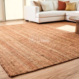 May include: A large rectangular area rug featuring a natural tan and brown woven texture with a consistent herringbone pattern throughout. The rug is laid flat on a light-colored wooden floor, displaying a dense, fibrous weave and earthy tones.