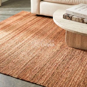 May include: A rectangular area rug featuring a woven herringbone pattern in shades of warm terracotta and natural tan. The rug has a textured, fibrous surface and is positioned on a floor partially covered by a light-colored coffee table and sofa.