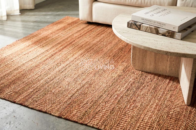 May include: A rectangular area rug featuring a woven herringbone pattern in shades of warm terracotta and natural tan. The rug has a textured, fibrous surface and is positioned on a floor partially covered by a light-colored coffee table and sofa.