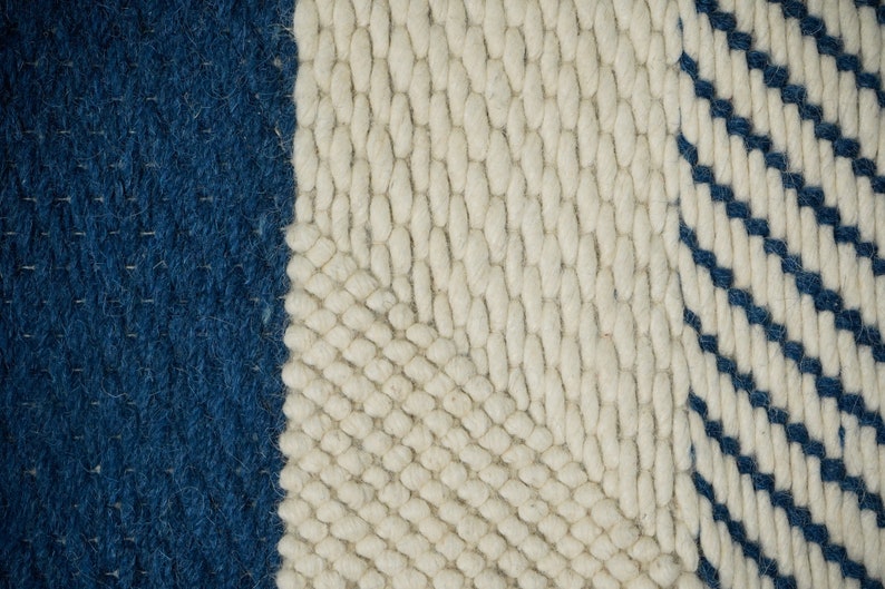May include: Close-up of a woven rug with a white background and blue stripes. The rug has a textured, hand-woven look.