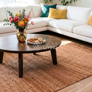 May include: A round dark brown wooden coffee table with four legs, featuring a glass vase filled with colorful flowers and a small plate of cookies on top, positioned on a large textured brown woven rug in front of a white sectional sofa.