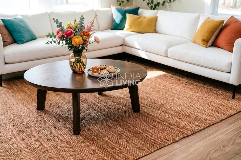 May include: A round dark brown wooden coffee table with four legs, featuring a glass vase filled with colorful flowers and a small plate of cookies on top, positioned on a large textured brown woven rug in front of a white sectional sofa.