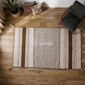 May include: A rectangular area rug featuring a central gray panel flanked by cream and dark brown striped sections with geometric patterns. The rug has tassels at each corner and displays the text INDIA LIVING in the center.