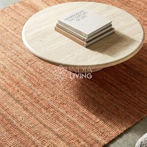 May include: A rectangular area rug with a woven herringbone pattern in shades of terracotta and orange. A round, light-colored wooden coffee table sits on the rug, topped with a stack of two books titled "HERRINGBONE THE TEXTURE OF LIFE".