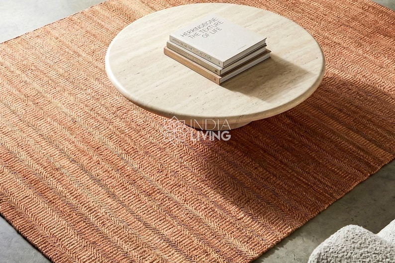 May include: A rectangular area rug with a woven herringbone pattern in shades of terracotta and orange. A round, light-colored wooden coffee table sits on the rug, topped with a stack of two books titled "HERRINGBONE THE TEXTURE OF LIFE".
