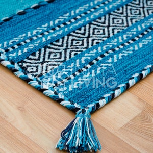 May include: A rectangular woven rug featuring a vibrant turquoise and blue color palette with a central band of black and white diamond patterns. The edges are finished with a braided border and a thick tassel in shades of blue and white.