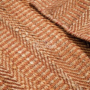 May include: A woven rug featuring a textured chevron pattern in shades of burnt orange and natural tan. The material appears to be a thick, fibrous natural fiber with a visible braided weave construction and a folded corner revealing the underside.