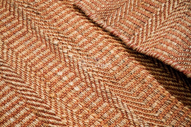 May include: A woven rug featuring a textured chevron pattern in shades of burnt orange and natural tan. The material appears to be a thick, fibrous natural fiber with a visible braided weave construction and a folded corner revealing the underside.