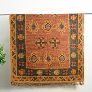 Puede incluir: Una alfombra rectangular con un patr&oacute;n geom&eacute;trico en tonos marrones, naranjas y amarillos. La alfombra tiene un borde con flecos.