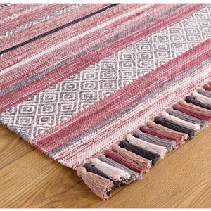 May include: A woven rug with a mix of stripes and diamond patterns in shades of red, gray, and white. The rug has a fringe of tassels in coordinating colors along one edge. The rug is on a wooden floor.