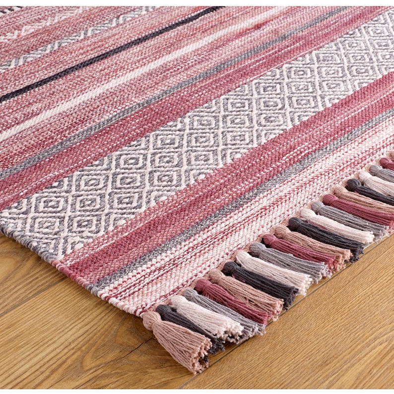 May include: A woven rug with a mix of stripes and diamond patterns in shades of red, gray, and white. The rug has a fringe of tassels in coordinating colors along one edge. The rug is on a wooden floor.