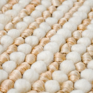 May include: Close-up of a woven rug with a textured surface. The rug is made of beige and white yarn and has a unique, bumpy texture.
