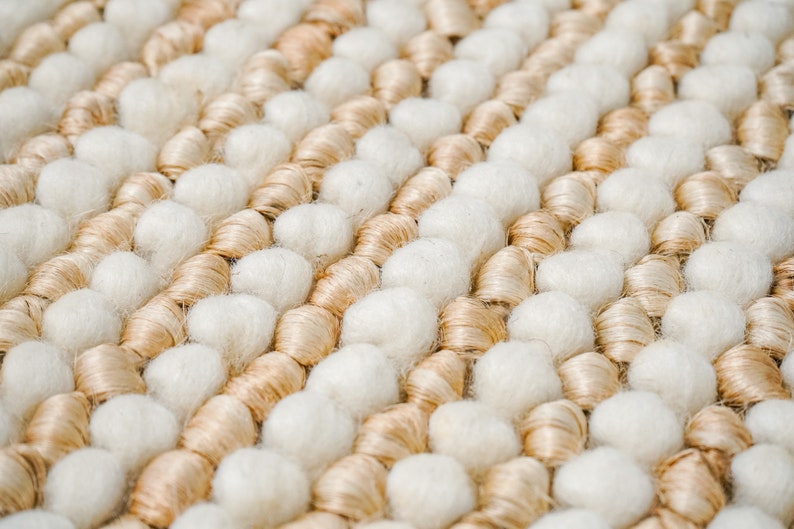 May include: Close-up of a woven rug with a textured surface. The rug is made of beige and white yarn and has a unique, bumpy texture.