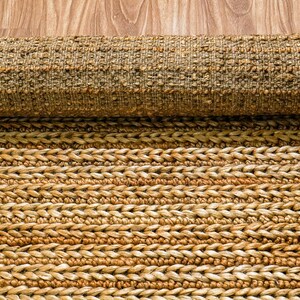 May include: A rolled-up, natural fiber rug with a textured, woven design. The rug is primarily tan and brown, with a braided pattern visible on the unrolled portion. The rug is made of a durable material, suitable for high-traffic areas.