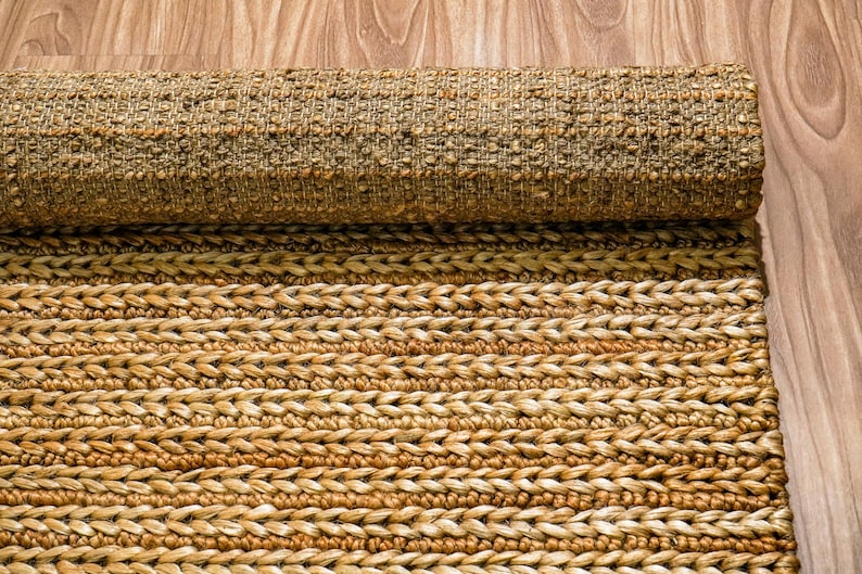 May include: A rolled-up, natural fiber rug with a textured, woven design. The rug is primarily tan and brown, with a braided pattern visible on the unrolled portion. The rug is made of a durable material, suitable for high-traffic areas.