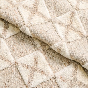 May include: Close-up of a beige and white woven rug with a checkered pattern. The rug has a soft, textured surface.
