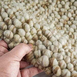 K&ouml;nnte beinhalten: Nahaufnahme eines beige-braunen Wollteppichs mit einer strukturierten, unebenen Oberfl&auml;che. Der Teppich besteht aus kleinen, runden Wollkugeln, die miteinander verwoben sind.