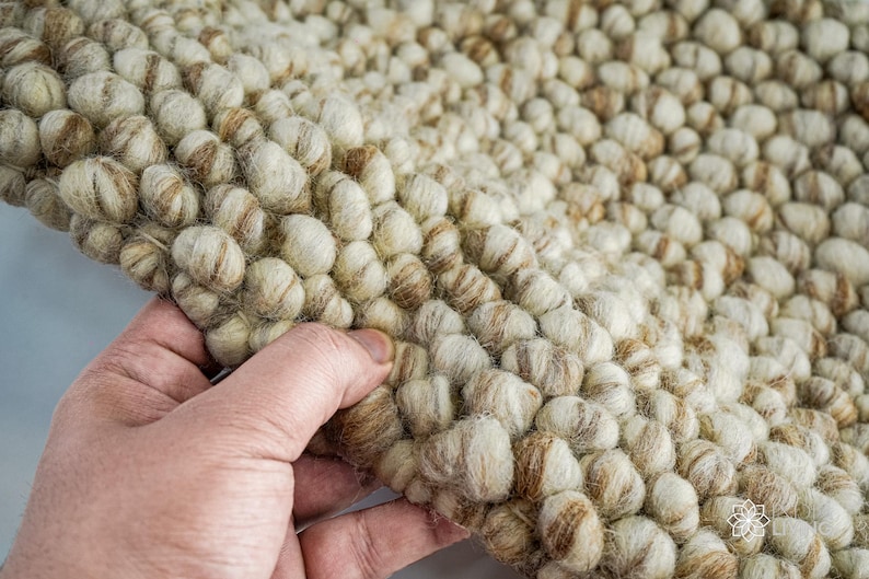K&ouml;nnte beinhalten: Nahaufnahme eines beige-braunen Wollteppichs mit einer strukturierten, unebenen Oberfl&auml;che. Der Teppich besteht aus kleinen, runden Wollkugeln, die miteinander verwoben sind.