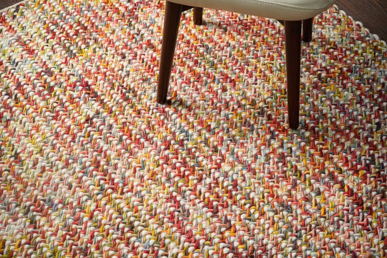 May include: A multi-colored braided area rug featuring a dense, woven texture with a vibrant mix of red, yellow, orange, blue, and white yarn strands. The rug has a thick, chunky construction and is partially covered by the legs of a chair.