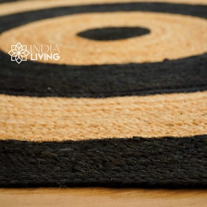 May include: A round jute rug with black and natural colored stripes. The rug has a concentric circle pattern. The rug is made of natural fibers and has a rustic look. The text "INDIA LIVING" is visible in the center of the rug.