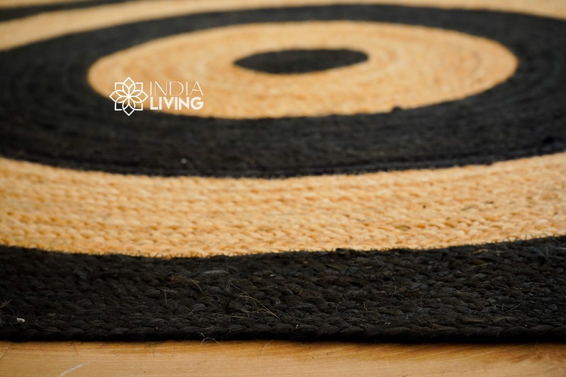 May include: A round jute rug with black and natural colored stripes. The rug has a concentric circle pattern. The rug is made of natural fibers and has a rustic look. The text "INDIA LIVING" is visible in the center of the rug.