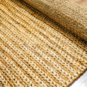 May include: A rolled-up, natural fiber rug with a woven, braided design. The rug is a warm, neutral color, with variations in tone. The texture is visible, and the rug appears to be made of jute or a similar material. The rug is partially rolled up.