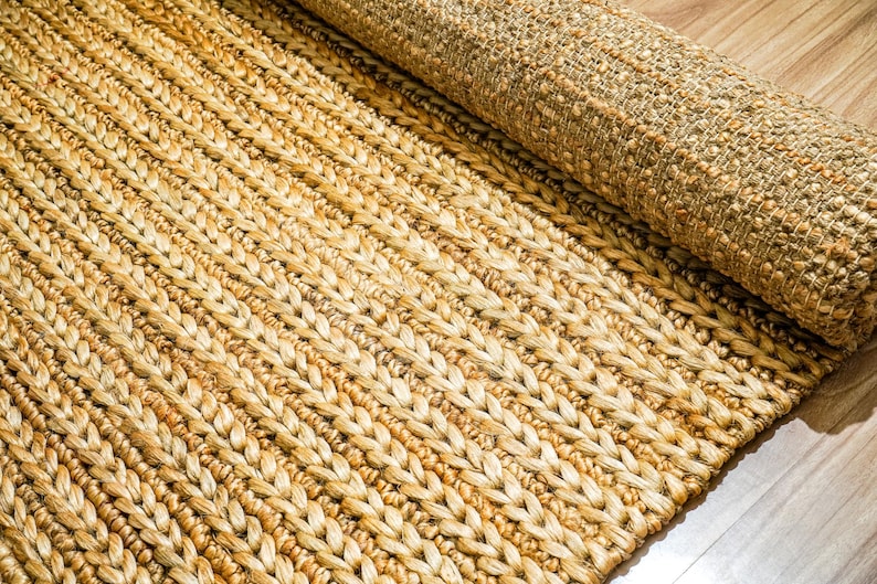 May include: A rolled-up, natural fiber rug with a woven, braided design. The rug is a warm, neutral color, with variations in tone. The texture is visible, and the rug appears to be made of jute or a similar material. The rug is partially rolled up.