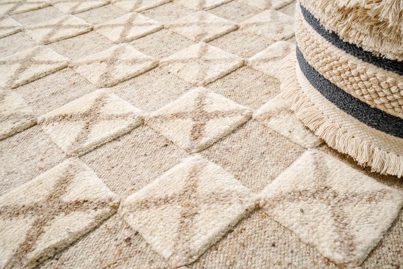May include: A close-up of a cream-colored rug with a geometric pattern of squares and diamonds. The rug has a subtle texture and a slightly distressed look.