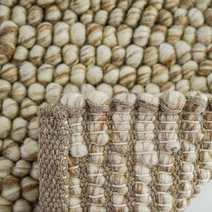 K&ouml;nnte beinhalten: Nahaufnahme eines runden, beige-braunen Teppichs mit einer strukturierten, gewebten Oberfl&auml;che. Der Teppich besteht aus kleinen, runden Garnkugeln.