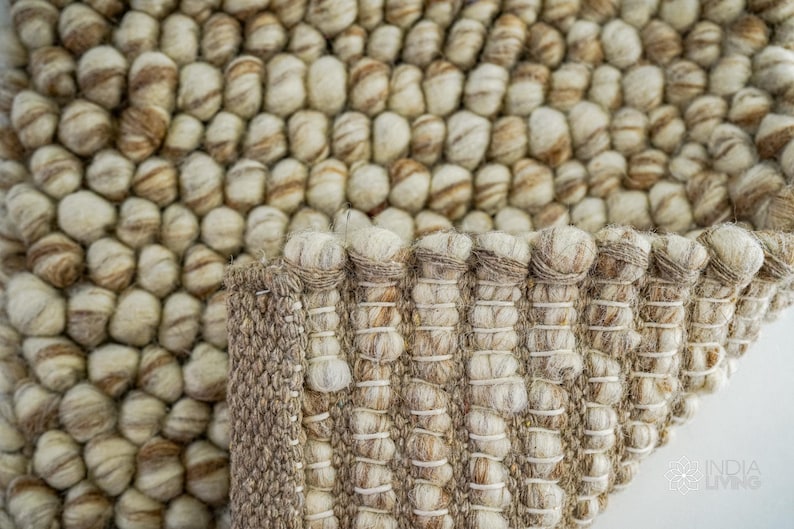 K&ouml;nnte beinhalten: Nahaufnahme eines runden, beige-braunen Teppichs mit einer strukturierten, gewebten Oberfl&auml;che. Der Teppich besteht aus kleinen, runden Garnkugeln.