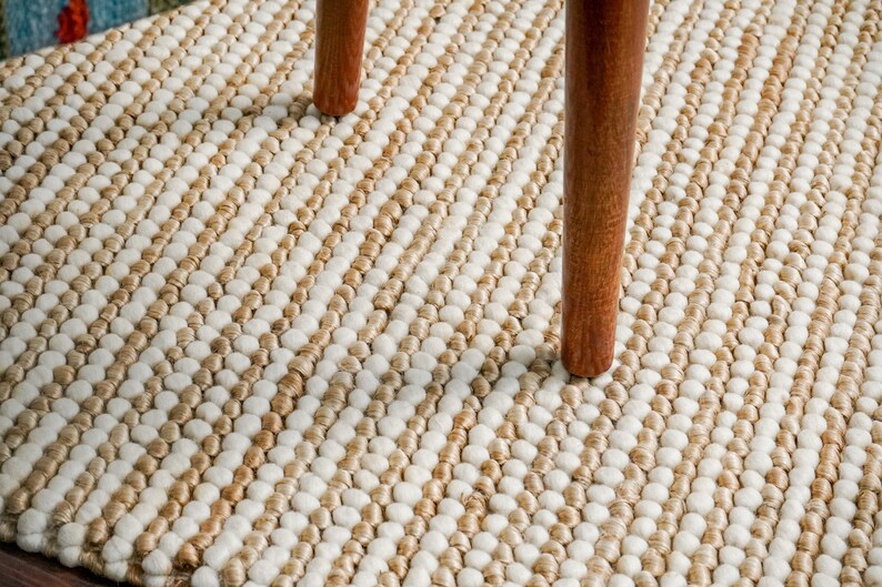 May include: Close-up view of a textured rug with a striped pattern in beige and off-white. The rug is made of small, round, looped yarn creating a nubby texture.  Two wooden chair legs are visible on the rug's surface, showcasing its durability and suitability for high-traffic areas. The rug's color palette is neutral and earthy, making it a versatile addition to various home decor styles.