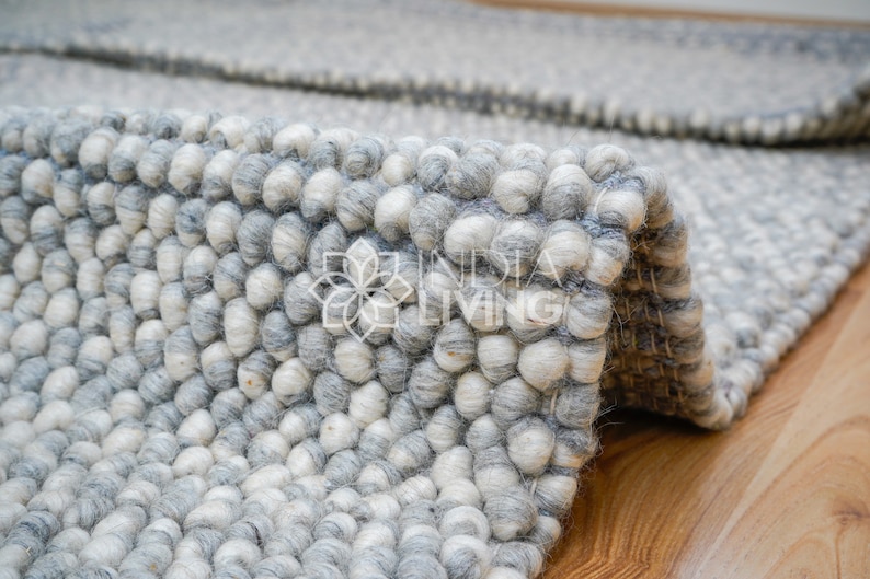 May include: Close-up of a gray and white wool rug with a textured, bumpy surface. The rug is made of small, round balls of wool that are tightly woven together.