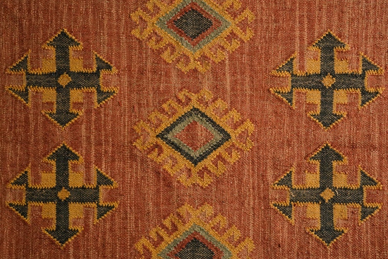 Puede incluir: Primer plano de una alfombra de color rojo, naranja y marr&oacute;n con un dise&ntilde;o geom&eacute;trico. La alfombra tiene una textura tejida y presenta un patr&oacute;n repetitivo de diamantes y cruces.