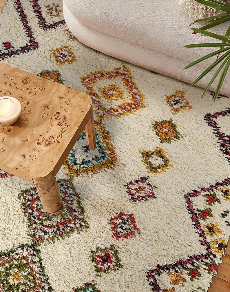 Op de afbeelding: Een wit shag-kleed met een kleurrijk geometrisch patroon. Het kleed heeft een ruitpatroon met rode, gele, groene en blauwe accenten. Het kleed wordt getoond met een houten tafel en een witte bank.