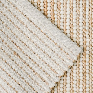 May include: Close-up of a cream-colored woven rug with a textured, bumpy surface. The rug has a striped pattern with alternating rows of small, round bumps and flat woven strands.