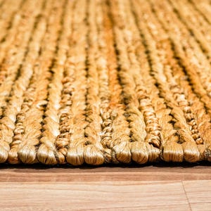 May include: Close-up of a woven jute rug with a natural, golden-brown color. The rug has a textured surface with a braided pattern, creating a rustic and inviting aesthetic. The rug is placed on a wooden floor.
