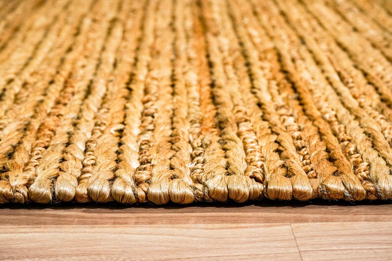 May include: Close-up of a woven jute rug with a natural, golden-brown color. The rug has a textured surface with a braided pattern, creating a rustic and inviting aesthetic. The rug is placed on a wooden floor.