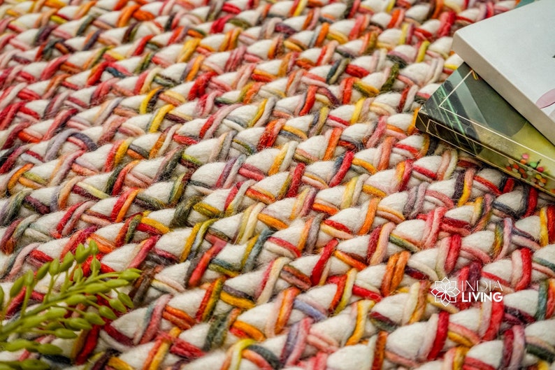 May include: A thick, hand-woven rug featuring a chunky braided texture. The design consists of cream-colored yarn interwoven with vibrant strands of red, orange, yellow, and dark green. The logo INDIA LIVING is visible in the bottom right corner.
