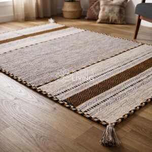 May include: A rectangular area rug featuring a central gray panel flanked by cream, tan, and dark brown striped sections with geometric patterns. The rug has a braided border and tassels at the corners. The text "INDUS LIVING" is visible in the center.