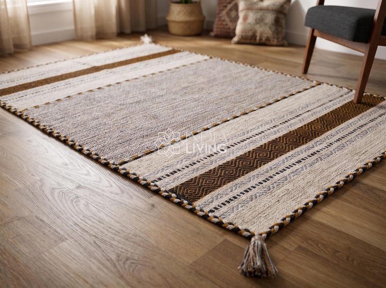 May include: A rectangular area rug featuring a central gray panel flanked by cream, tan, and dark brown striped sections with geometric patterns. The rug has a braided border and tassels at the corners. The text "INDUS LIVING" is visible in the center.