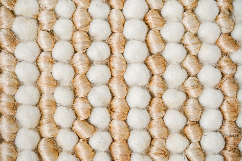 May include: Close-up of a woven rug with a pattern of beige and white yarn balls.