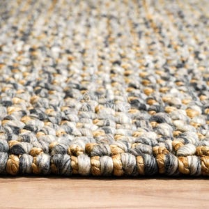 Puede incluir: Una alfombra gruesa tejida a mano con una textura trenzada y voluminosa. La alfombra est&aacute; compuesta por una mezcla de hilos en tonos gris carb&oacute;n, gris claro, crema y marr&oacute;n dorado c&aacute;lido, creando un aspecto variado y multicolor en toda la pieza.