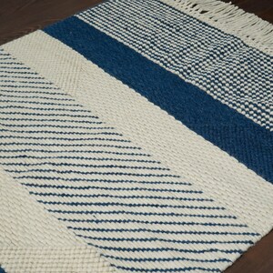May include: A white and blue woven rug with a geometric pattern. The rug has a fringe border and is made of natural fibers.